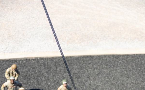 A military soldier in full combat uniform and helmet is rappelling down a rope from an elevated position, viewed from above. A group of approximately 10-12 soldiers in camouflage uniforms and helmets are gathered on the ground below, observing the descent. This appears to be a military rappelling or airborne training exercise.