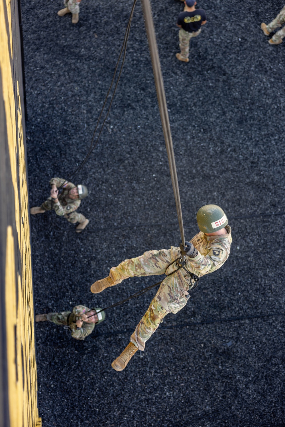 An aerial photograph taken from above shows a military personnel rappelling down a rope from an elevated structure, likely a tower or building used for training purposes. The soldier is wearing full camouflage uniform, a green helmet with white labeling, and is secured by a harness system. Several other uniformed personnel are visible on the ground below, observing or awaiting their turn.