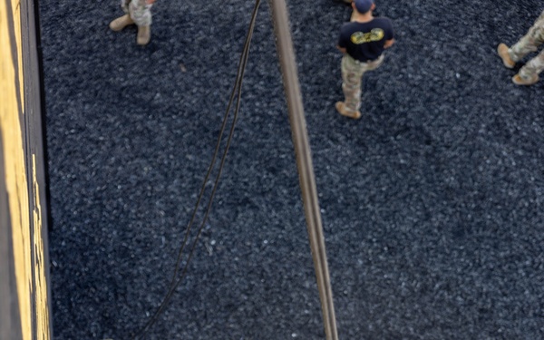 An aerial photograph taken from above shows a military personnel rappelling down a rope from an elevated structure, likely a tower or building used for training purposes. The soldier is wearing full camouflage uniform, a green helmet with white labeling, and is secured by a harness system. Several other uniformed personnel are visible on the ground below, observing or awaiting their turn.