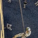 An aerial photograph taken from above shows a military personnel rappelling down a rope from an elevated structure, likely a tower or building used for training purposes. The soldier is wearing full camouflage uniform, a green helmet with white labeling, and is secured by a harness system. Several other uniformed personnel are visible on the ground below, observing or awaiting their turn.