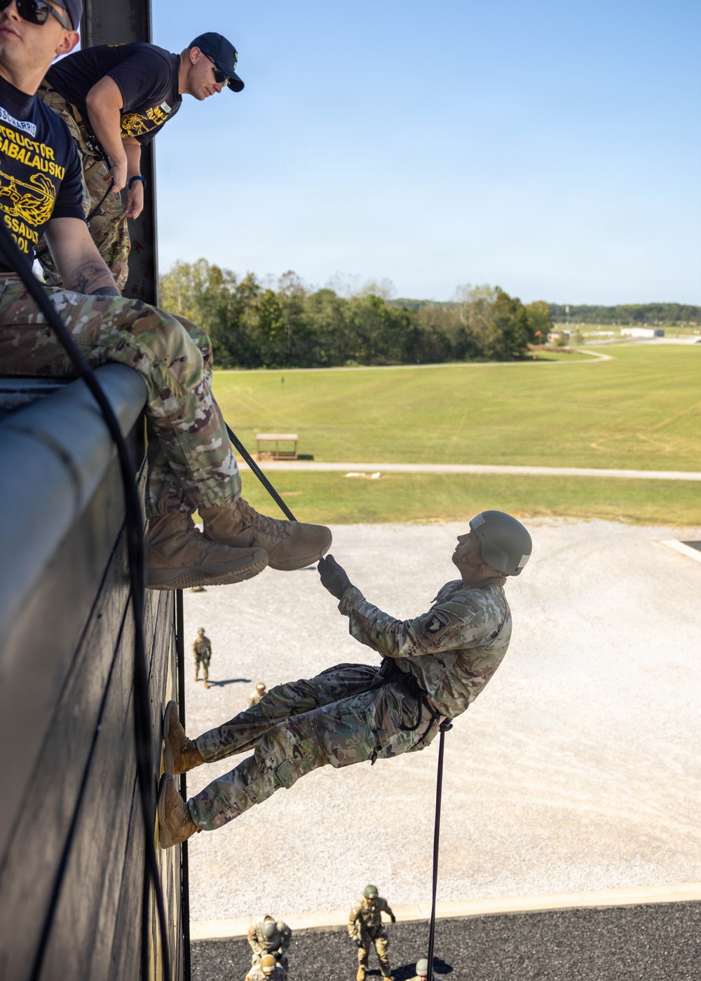 Military personnel are conducting a rappelling training exercise from an elevated structure, likely a rappel tower. An instructor wearing a navy blue shirt with yellow text reading 'INSTRUCTOR' and partial text 'SABALAUSKI' and 'SSAULT SCHOOL' oversees a soldier in full camouflage uniform and helmet descending the wall on a rope. Additional personnel are visible on the ground below, suggesting this is a supervised training event.