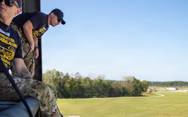 Military personnel are conducting a rappelling training exercise from an elevated structure, likely a rappel tower. An instructor wearing a navy blue shirt with yellow text reading 'INSTRUCTOR' and partial text 'SABALAUSKI' and 'SSAULT SCHOOL' oversees a soldier in full camouflage uniform and helmet descending the wall on a rope. Additional personnel are visible on the ground below, suggesting this is a supervised training event.