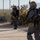 56th CRD Dugway VALEX 2026 Two heavily equipped military or tactical personnel are moving along a paved path adjacent to a chain-link fence in an arid, desert-like environment. Both individuals are wearing gas masks, tactical helmets, MOLLE plate carriers, and are carrying what appear to be rebreather or SCBA units on their backs. They are armed with rifles and appear to be conducting a tactical exercise or operation.