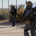 Two heavily equipped military or tactical personnel are moving along a paved path adjacent to a chain-link fence in an arid, desert-like environment. Both individuals are wearing gas masks, tactical helmets, MOLLE plate carriers, and are carrying what appear to be rebreather or SCBA units on their backs. They are armed with rifles and appear to be conducting a tactical exercise or operation.