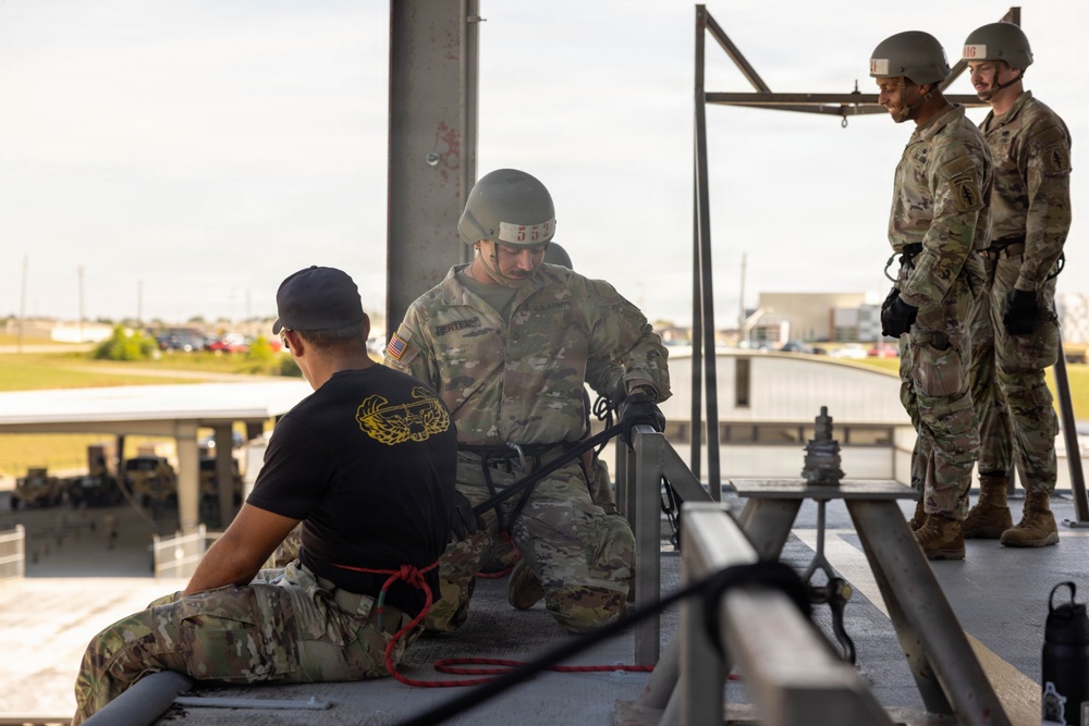 Military personnel are engaged in what appears to be rappelling or rope equipment training on an elevated platform structure. One soldier in OCP (Operational Camouflage Pattern) uniform and helmet is kneeling and handling harness/rope equipment, while an instructor in a black t-shirt observes. Two additional soldiers in full OCP uniforms and helmets stand nearby, apparently waiting or observing the training exercise.