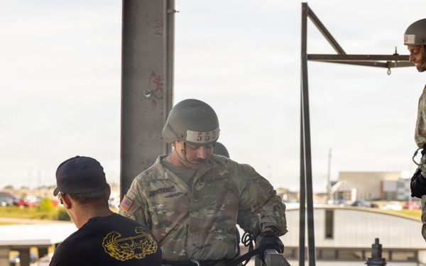 Military personnel are engaged in what appears to be rappelling or rope equipment training on an elevated platform structure. One soldier in OCP (Operational Camouflage Pattern) uniform and helmet is kneeling and handling harness/rope equipment, while an instructor in a black t-shirt observes. Two additional soldiers in full OCP uniforms and helmets stand nearby, apparently waiting or observing the training exercise.