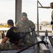 Military personnel are engaged in what appears to be rappelling or rope equipment training on an elevated platform structure. One soldier in OCP (Operational Camouflage Pattern) uniform and helmet is kneeling and handling harness/rope equipment, while an instructor in a black t-shirt observes. Two additional soldiers in full OCP uniforms and helmets stand nearby, apparently waiting or observing the training exercise.