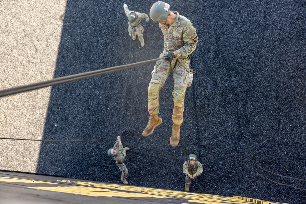 A top-down aerial photograph captures multiple U.S. Army soldiers rappelling down a vertical surface, likely a training tower. The perspective is shot from above, showing soldiers at various stages of their descent along ropes against a dark textured surface. The closest soldier is clearly visible in full OCP (Operational Camouflage Pattern) uniform with a helmet and gloves.