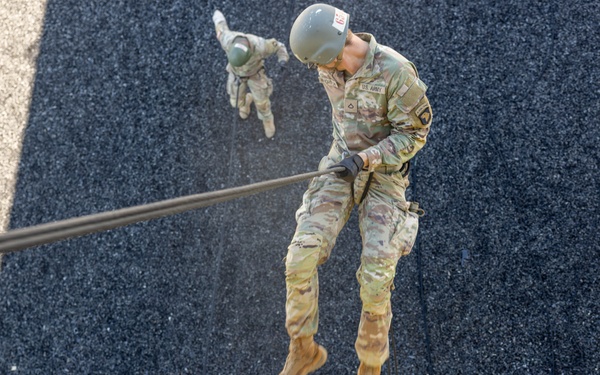 A top-down aerial photograph captures multiple U.S. Army soldiers rappelling down a vertical surface, likely a training tower. The perspective is shot from above, showing soldiers at various stages of their descent along ropes against a dark textured surface. The closest soldier is clearly visible in full OCP (Operational Camouflage Pattern) uniform with a helmet and gloves.