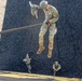 A top-down aerial photograph captures multiple U.S. Army soldiers rappelling down a vertical surface, likely a training tower. The perspective is shot from above, showing soldiers at various stages of their descent along ropes against a dark textured surface. The closest soldier is clearly visible in full OCP (Operational Camouflage Pattern) uniform with a helmet and gloves.
