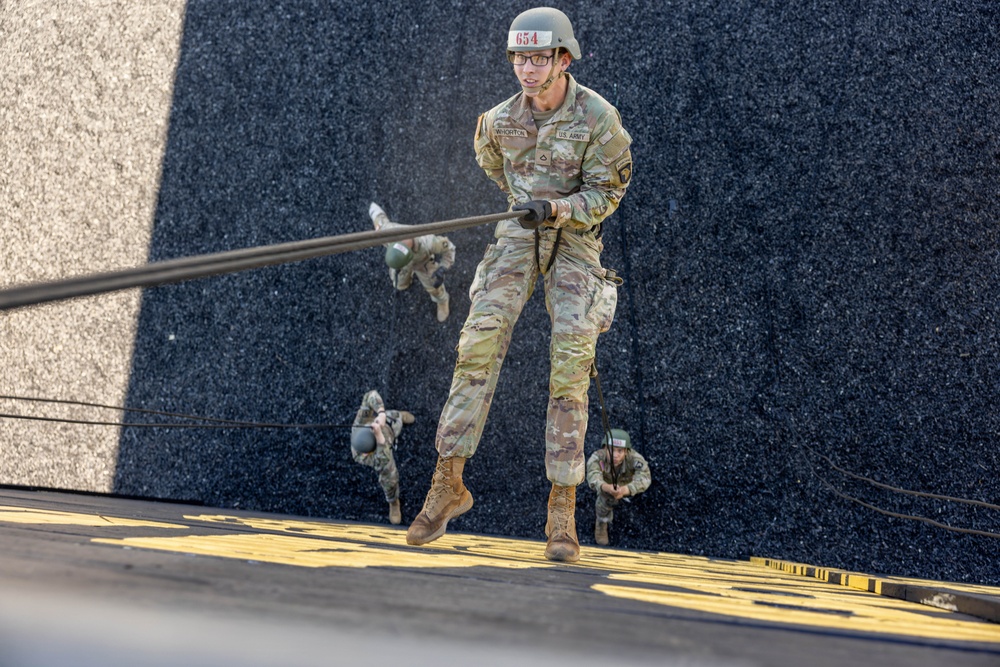 A U.S. Army soldier wearing helmet number 654 and identified by the name tape 'WHORTON' is performing a rappelling exercise down a steep inclined surface. The soldier is in full OCP (Operational Camouflage Pattern) uniform, gripping a rope while descending. Two additional soldiers in similar uniforms are visible in the background on the same inclined surface.
