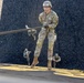A U.S. Army soldier wearing helmet number 654 and identified by the name tape 'WHORTON' is performing a rappelling exercise down a steep inclined surface. The soldier is in full OCP (Operational Camouflage Pattern) uniform, gripping a rope while descending. Two additional soldiers in similar uniforms are visible in the background on the same inclined surface.