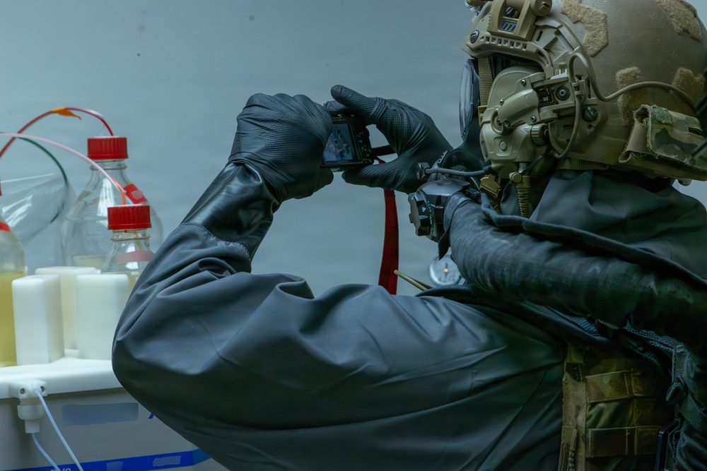A person wearing full tactical/protective gear including a military-style helmet, gas mask, and black hazmat-style suit is photographing laboratory equipment with a small digital camera. The scene appears to be a controlled environment inspection or documentation of a chemical laboratory setting. The individual is equipped with what appears to be CBRN (Chemical, Biological, Radiological, Nuclear) protective equipment.