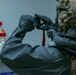 A person wearing full tactical/protective gear including a military-style helmet, gas mask, and black hazmat-style suit is photographing laboratory equipment with a small digital camera. The scene appears to be a controlled environment inspection or documentation of a chemical laboratory setting. The individual is equipped with what appears to be CBRN (Chemical, Biological, Radiological, Nuclear) protective equipment.