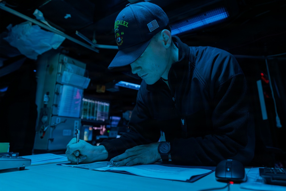 A U.S. Navy sailor is writing or reviewing documents at a workstation aboard what appears to be a naval vessel, illuminated by blue ambient lighting typical of a ship's combat information center or bridge. The sailor is wearing a cap with 'GONZALEZ' and the number '66' visible, along with a U.S. flag patch, indicating assignment to USS Gonzalez (DDG-66). The low-light blue environment and background displays suggest a nighttime operational setting.