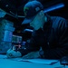 A U.S. Navy sailor is writing or reviewing documents at a workstation aboard what appears to be a naval vessel, illuminated by blue ambient lighting typical of a ship's combat information center or bridge. The sailor is wearing a cap with 'GONZALEZ' and the number '66' visible, along with a U.S. flag patch, indicating assignment to USS Gonzalez (DDG-66). The low-light blue environment and background displays suggest a nighttime operational setting.