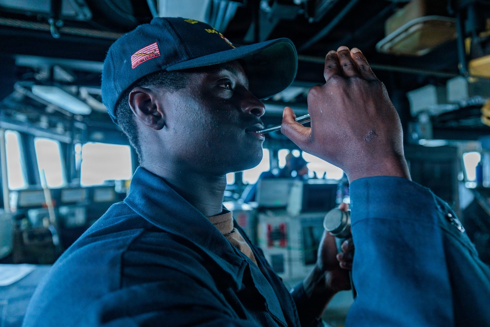 A U.S. Navy sailor wearing a dark blue naval cap with a red emblem is using a navigation instrument, likely a bearing circle or pelorus, on what appears to be the bridge of a naval vessel. The sailor is in uniform and appears to be taking a navigational sighting or bearing reading. The bridge environment includes various control panels, monitors, and equipment visible in the background.