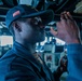 A U.S. Navy sailor wearing a dark blue naval cap with a red emblem is using a navigation instrument, likely a bearing circle or pelorus, on what appears to be the bridge of a naval vessel. The sailor is in uniform and appears to be taking a navigational sighting or bearing reading. The bridge environment includes various control panels, monitors, and equipment visible in the background.