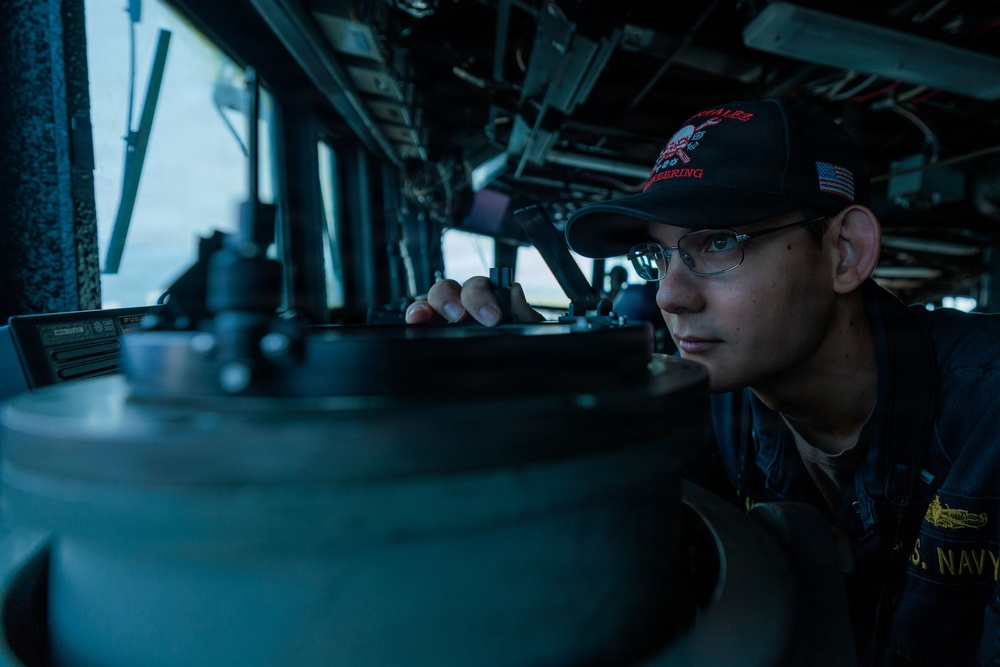 A U.S. Navy sailor is stationed on the bridge of a naval vessel, peering through or over a navigational instrument, likely a gyrocompass or pelorus. The sailor is wearing a dark ball cap with an American flag patch and what appears to be a ship-specific insignia, along with a U.S. Navy uniform. The image is taken from a low angle inside the bridge, with ocean visible through the windows in the background.