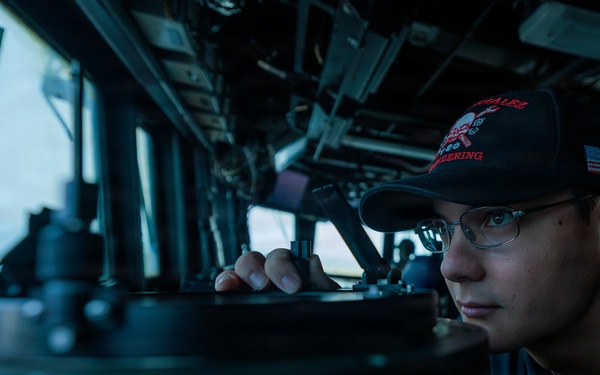 A U.S. Navy sailor is stationed on the bridge of a naval vessel, peering through or over a navigational instrument, likely a gyrocompass or pelorus. The sailor is wearing a dark ball cap with an American flag patch and what appears to be a ship-specific insignia, along with a U.S. Navy uniform. The image is taken from a low angle inside the bridge, with ocean visible through the windows in the background.