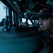 A U.S. Navy sailor is stationed on the bridge of a naval vessel, peering through or over a navigational instrument, likely a gyrocompass or pelorus. The sailor is wearing a dark ball cap with an American flag patch and what appears to be a ship-specific insignia, along with a U.S. Navy uniform. The image is taken from a low angle inside the bridge, with ocean visible through the windows in the background.