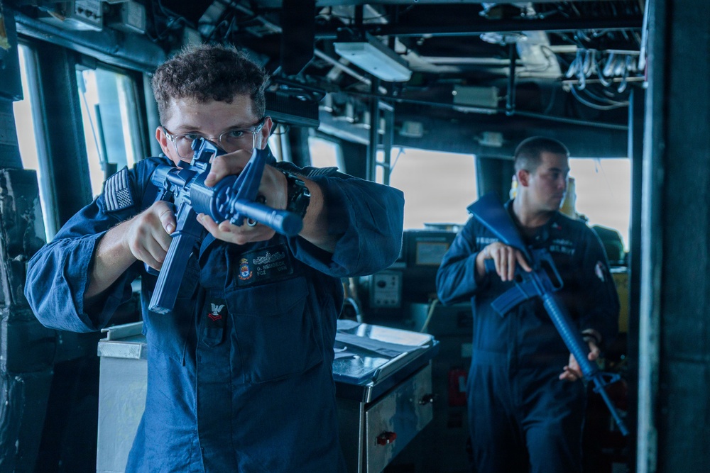 Two U.S. Navy sailors in blue coverall uniforms are conducting what appears to be a security drill or training exercise aboard a naval vessel's bridge or interior compartment. Both personnel are holding rifles in a ready or aiming position. The foreground sailor wears safety glasses and has visible rank insignia and a U.S. flag patch on the sleeve.