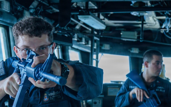 Two U.S. Navy sailors in blue coverall uniforms are conducting what appears to be a security drill or training exercise aboard a naval vessel's bridge or interior compartment. Both personnel are holding rifles in a ready or aiming position. The foreground sailor wears safety glasses and has visible rank insignia and a U.S. flag patch on the sleeve.