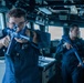 Two U.S. Navy sailors in blue coverall uniforms are conducting what appears to be a security drill or training exercise aboard a naval vessel's bridge or interior compartment. Both personnel are holding rifles in a ready or aiming position. The foreground sailor wears safety glasses and has visible rank insignia and a U.S. flag patch on the sleeve.