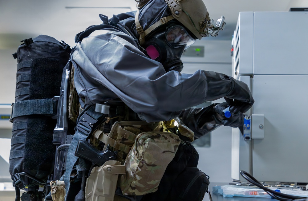 A person in full CBRN (Chemical, Biological, Radiological, Nuclear) protective gear is working at an electrical or equipment panel in an indoor facility. The individual is wearing a gas mask, protective oversuit, tactical helmet, and military-style load-bearing equipment. The scene appears to be a training exercise or operational response scenario.