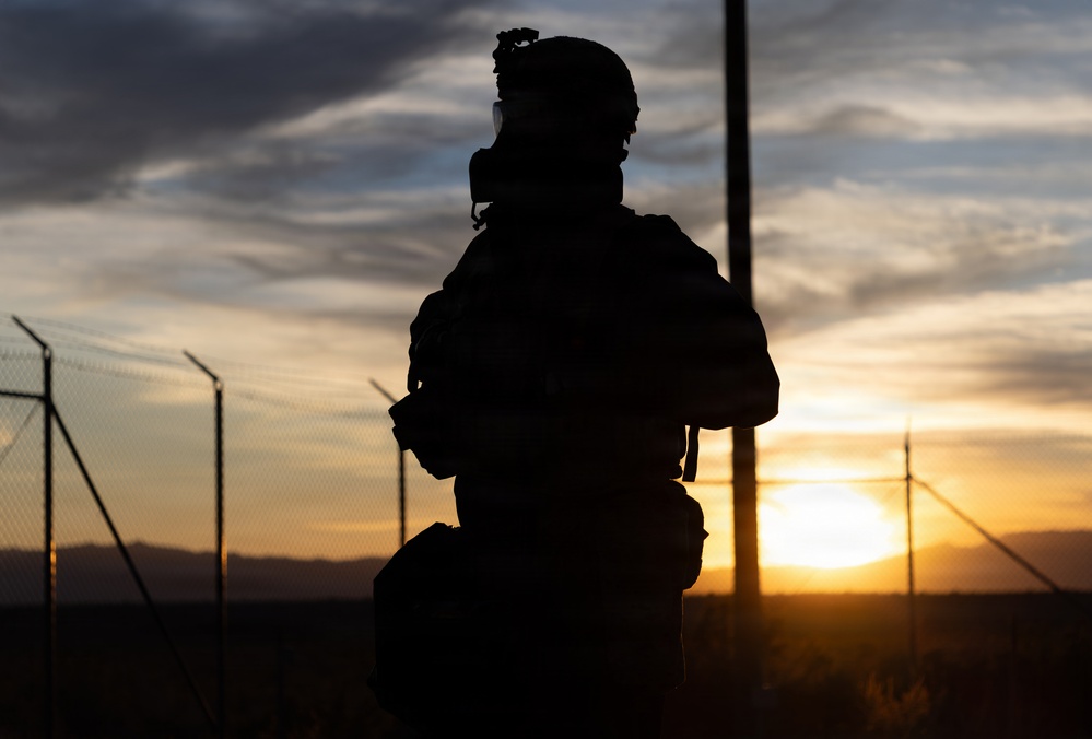 A silhouetted figure in full tactical/military gear is photographed against a dramatic sunset backdrop. The subject appears to be wearing a helmet with mounted equipment, a gas mask, body armor, and a backpack. A chain-link fence and utility poles are visible in the background, suggesting a secured perimeter location.