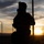 56th CRD Dugway VALEX 2026 A silhouetted figure in full tactical/military gear is photographed against a dramatic sunset backdrop. The subject appears to be wearing a helmet with mounted equipment, a gas mask, body armor, and a backpack. A chain-link fence and utility poles are visible in the background, suggesting a secured perimeter location.