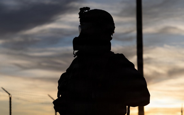 A silhouetted figure in full tactical/military gear is photographed against a dramatic sunset backdrop. The subject appears to be wearing a helmet with mounted equipment, a gas mask, body armor, and a backpack. A chain-link fence and utility poles are visible in the background, suggesting a secured perimeter location.