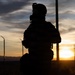 A silhouetted figure in full tactical/military gear is photographed against a dramatic sunset backdrop. The subject appears to be wearing a helmet with mounted equipment, a gas mask, body armor, and a backpack. A chain-link fence and utility poles are visible in the background, suggesting a secured perimeter location.