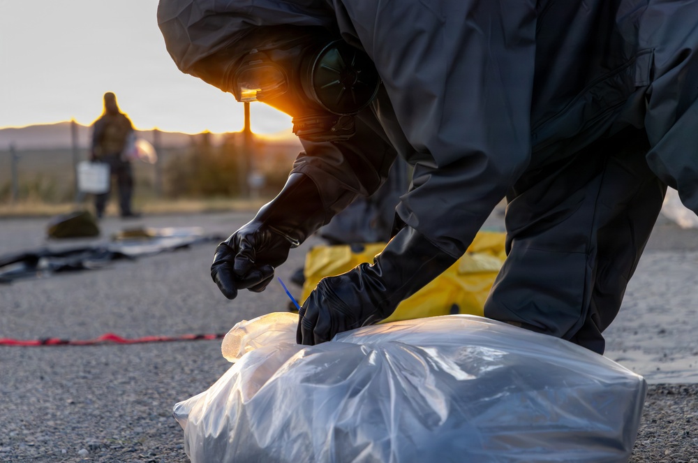 A person wearing a full hazmat/CBRN protective suit, gas mask, and black rubber gloves is kneeling on pavement and collecting or sampling material from a clear plastic bag. A second suited individual is visible in the background near what appears to be a perimeter marker. The scene appears to take place outdoors at dusk or dawn, suggesting an emergency response or training exercise.