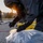56th CRD Dugway VALEX 2026 A person wearing a full hazmat/CBRN protective suit, gas mask, and black rubber gloves is kneeling on pavement and collecting or sampling material from a clear plastic bag. A second suited individual is visible in the background near what appears to be a perimeter marker. The scene appears to take place outdoors at dusk or dawn, suggesting an emergency response or training exercise.