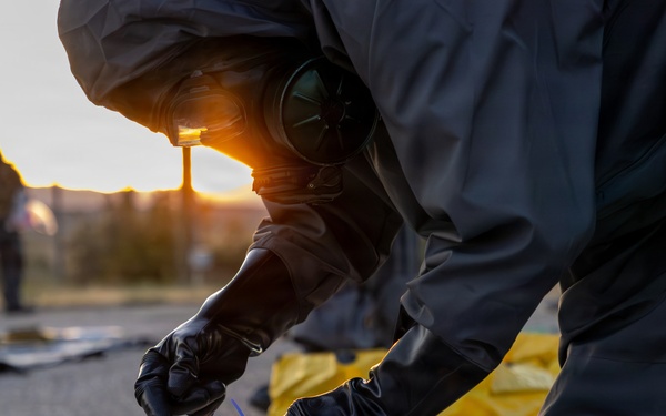 A person wearing a full hazmat/CBRN protective suit, gas mask, and black rubber gloves is kneeling on pavement and collecting or sampling material from a clear plastic bag. A second suited individual is visible in the background near what appears to be a perimeter marker. The scene appears to take place outdoors at dusk or dawn, suggesting an emergency response or training exercise.