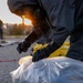 A person wearing a full hazmat/CBRN protective suit, gas mask, and black rubber gloves is kneeling on pavement and collecting or sampling material from a clear plastic bag. A second suited individual is visible in the background near what appears to be a perimeter marker. The scene appears to take place outdoors at dusk or dawn, suggesting an emergency response or training exercise.