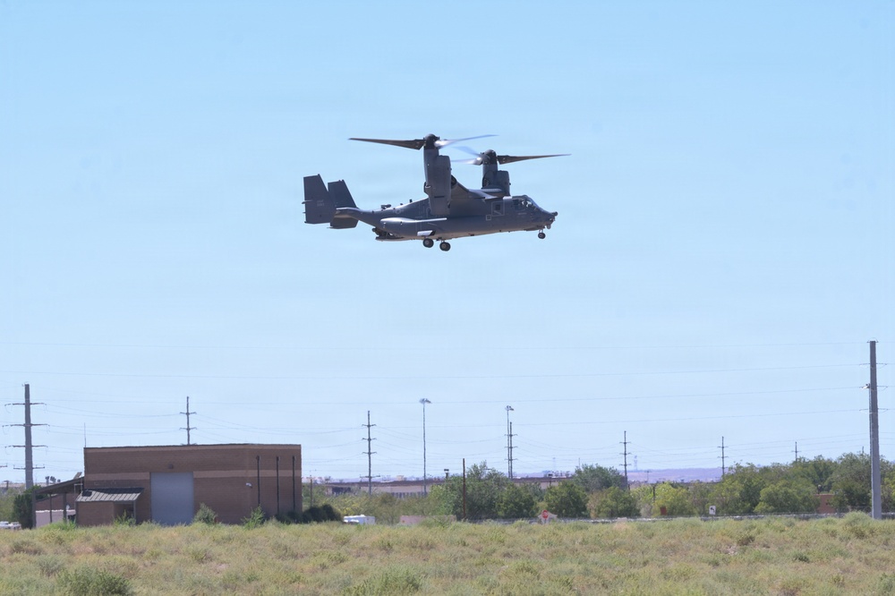A military tiltrotor aircraft, identifiable as a V-22 Osprey, is captured in low-altitude flight over a semi-arid landscape. The aircraft appears to be in helicopter mode with rotors tilted upward, suggesting it is either landing or taking off. The surrounding environment includes utility infrastructure, sparse vegetation, and low-rise buildings consistent with a military base or airfield setting.