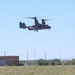 A military tiltrotor aircraft, identifiable as a V-22 Osprey, is captured in low-altitude flight over a semi-arid landscape. The aircraft appears to be in helicopter mode with rotors tilted upward, suggesting it is either landing or taking off. The surrounding environment includes utility infrastructure, sparse vegetation, and low-rise buildings consistent with a military base or airfield setting.