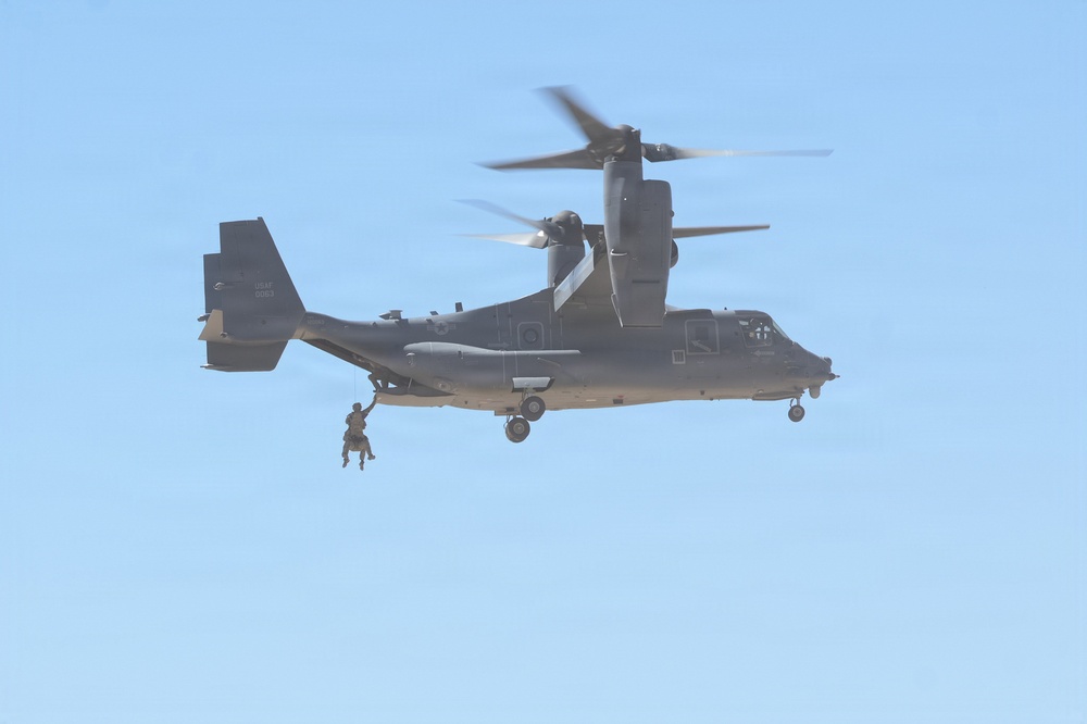 A U.S. Air Force CV-22 Osprey tiltrotor aircraft is hovering in flight against a clear blue sky. A personnel figure is visible rappelling or being extracted via rope from the underside/side of the aircraft. The rotors are in motion, visible as blurred arcs, indicating active flight.