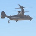 A U.S. Air Force CV-22 Osprey tiltrotor aircraft is hovering in flight against a clear blue sky. A personnel figure is visible rappelling or being extracted via rope from the underside/side of the aircraft. The rotors are in motion, visible as blurred arcs, indicating active flight.