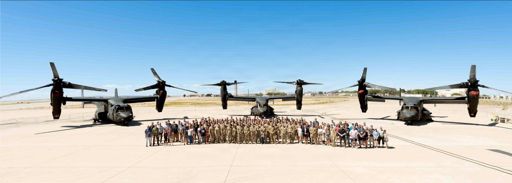 A large group of military personnel and civilians pose for a formal group photograph on a military airfield tarmac. Behind them are three tiltrotor aircraft, identifiable as CV-22 or V-22 Osprey variants, arranged symmetrically. The setting appears to be a sunny day at a desert or arid-climate military installation.