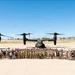 A large group of military personnel and civilians pose for a formal group photograph on a military airfield tarmac. Behind them are three tiltrotor aircraft, identifiable as CV-22 or V-22 Osprey variants, arranged symmetrically. The setting appears to be a sunny day at a desert or arid-climate military installation.