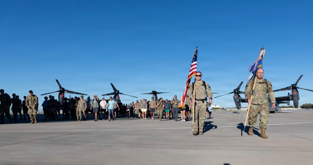 A military formation march or ruck event is taking place on an airfield tarmac, led by two soldiers in camouflage uniforms carrying the American flag and a unit guidon. A large group of participants, including both uniformed military personnel and civilians in casual attire, follow behind. Large tiltrotor aircraft, consistent with V-22 Osprey variants, are visible in the background.
