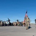 A military formation march or ruck event is taking place on an airfield tarmac, led by two soldiers in camouflage uniforms carrying the American flag and a unit guidon. A large group of participants, including both uniformed military personnel and civilians in casual attire, follow behind. Large tiltrotor aircraft, consistent with V-22 Osprey variants, are visible in the background.