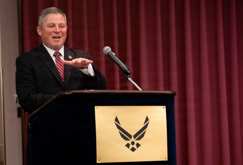 A man in a dark suit with a red striped tie is speaking at a podium bearing the United States Air Force emblem on a gold placard. He appears to be delivering a speech or address at an official Air Force event, gesturing with his right hand. The setting features a deep red curtain backdrop in what appears to be a formal venue or auditorium.