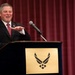 A man in a dark suit with a red striped tie is speaking at a podium bearing the United States Air Force emblem on a gold placard. He appears to be delivering a speech or address at an official Air Force event, gesturing with his right hand. The setting features a deep red curtain backdrop in what appears to be a formal venue or auditorium.