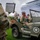 Military personnel are participating in a ceremonial event at a large football stadium. A soldier in the foreground holds a brass instrument (euphonium or baritone horn) with sheet music attached, while other soldiers ride in a vintage olive-drab military jeep labeled 'Colonel Saunders' with a white star emblem. The setting appears to be a pre-game or halftime military appreciation ceremony.