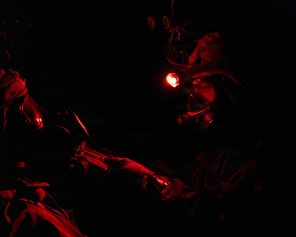 A low-light tactical scene illuminated by red light, showing at least one heavily equipped military or special operations personnel wearing a gas mask, tactical helmet with mounted equipment, and night vision device mounts. A firearm is visible being held in the foreground. The image is predominantly dark with red illumination as the primary light source.