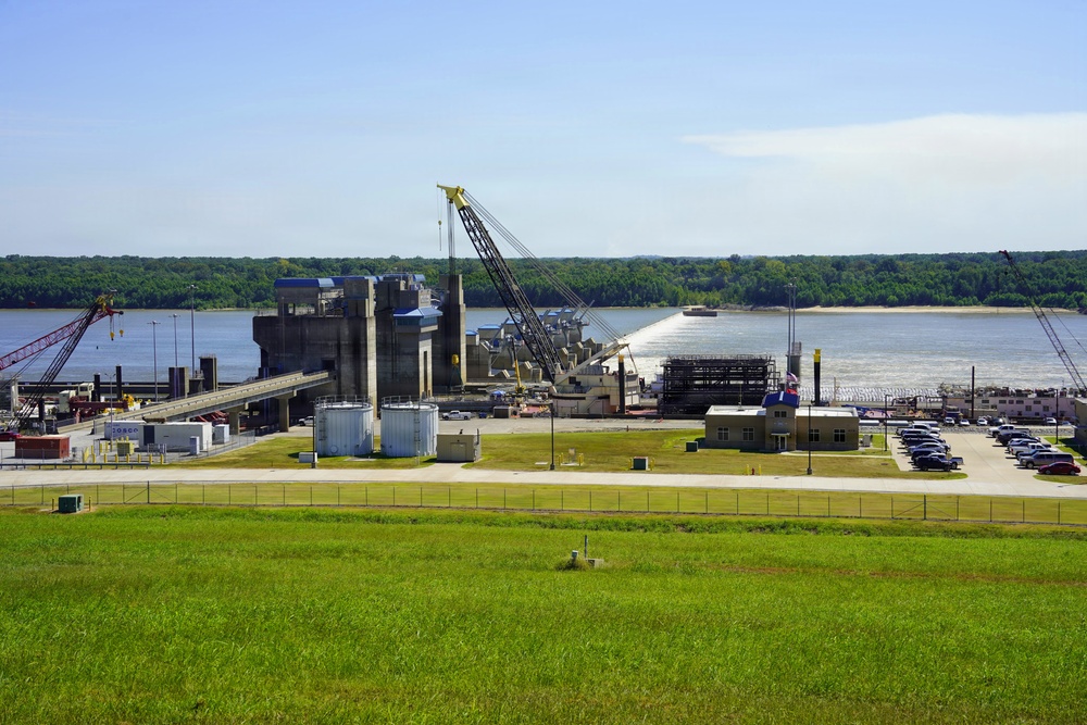 An aerial-perspective view of a river lock and dam facility, likely on a major U.S. inland waterway, showing active construction or maintenance operations. The facility includes concrete lock structures, cranes, storage tanks, and support buildings situated along a wide river with forested banks on the opposite shore. A parking area with numerous vehicles and what appears to be a barge on the river are also visible.